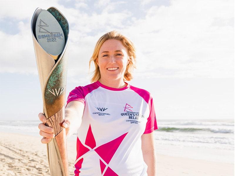 Blond female holding the Queen's baton at arms length standing on a sandy beach