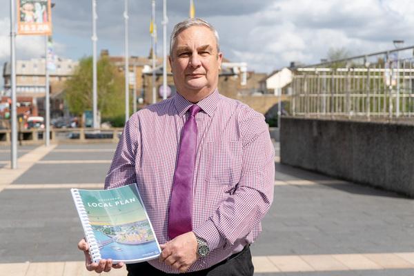 A man, Councillor Jjohn Burden standing outside, wearing a purple shirt and tie, holding a hardcopy ring bound draft local plan