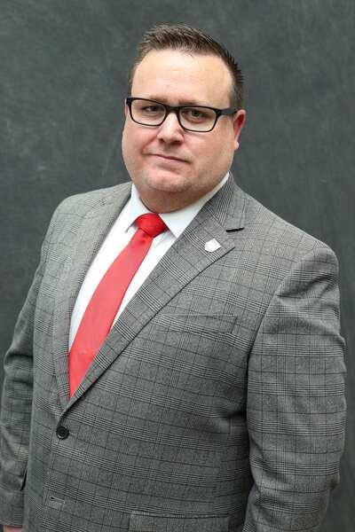 Portrait of man who is Councillor Shane Mochrie Cox, wearing a grey chequered blacker and a red tie.