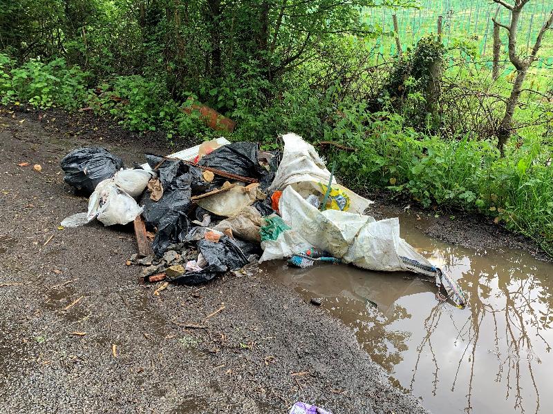 Mixed rubbish bags and rubbish dumped on a muddy field next to a puddle.