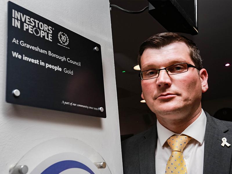 Male with dark hair, standing aside a dark plaque reading: investors in people at Gravesham Borough Council - we invest in people (gold)