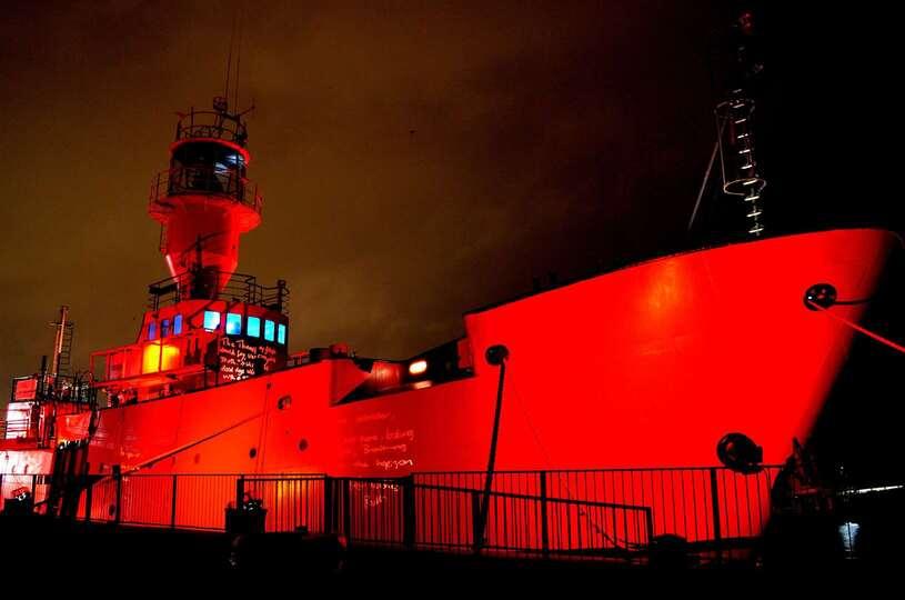 Red lighthouse vessel at night taken from the riverside, showing the front of the boat lit up.