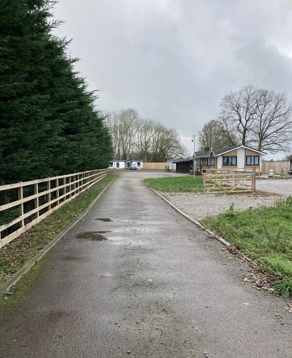 Looking down a road/path with a fence and tall conifer trees to the left and some buildings to the bottom right.