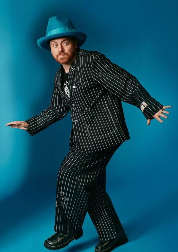 Main wearing a black pinstripe suite and a blue hat. He is stepping to the side and looking back. Against a teel blue background.