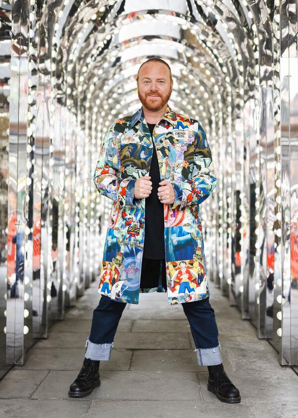 Leigh francis standing wearing a colourful long coat, with turned up dark denim jeans and boots, standing in an archway of mirrored lights.