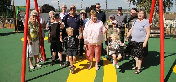 Group of adults and children sitting on and by the new swings in a play area.