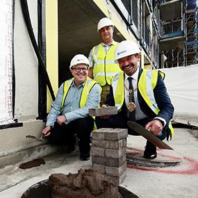 Three council officials wearing protective equipment, kneeling down to lay a brick on the housing development.