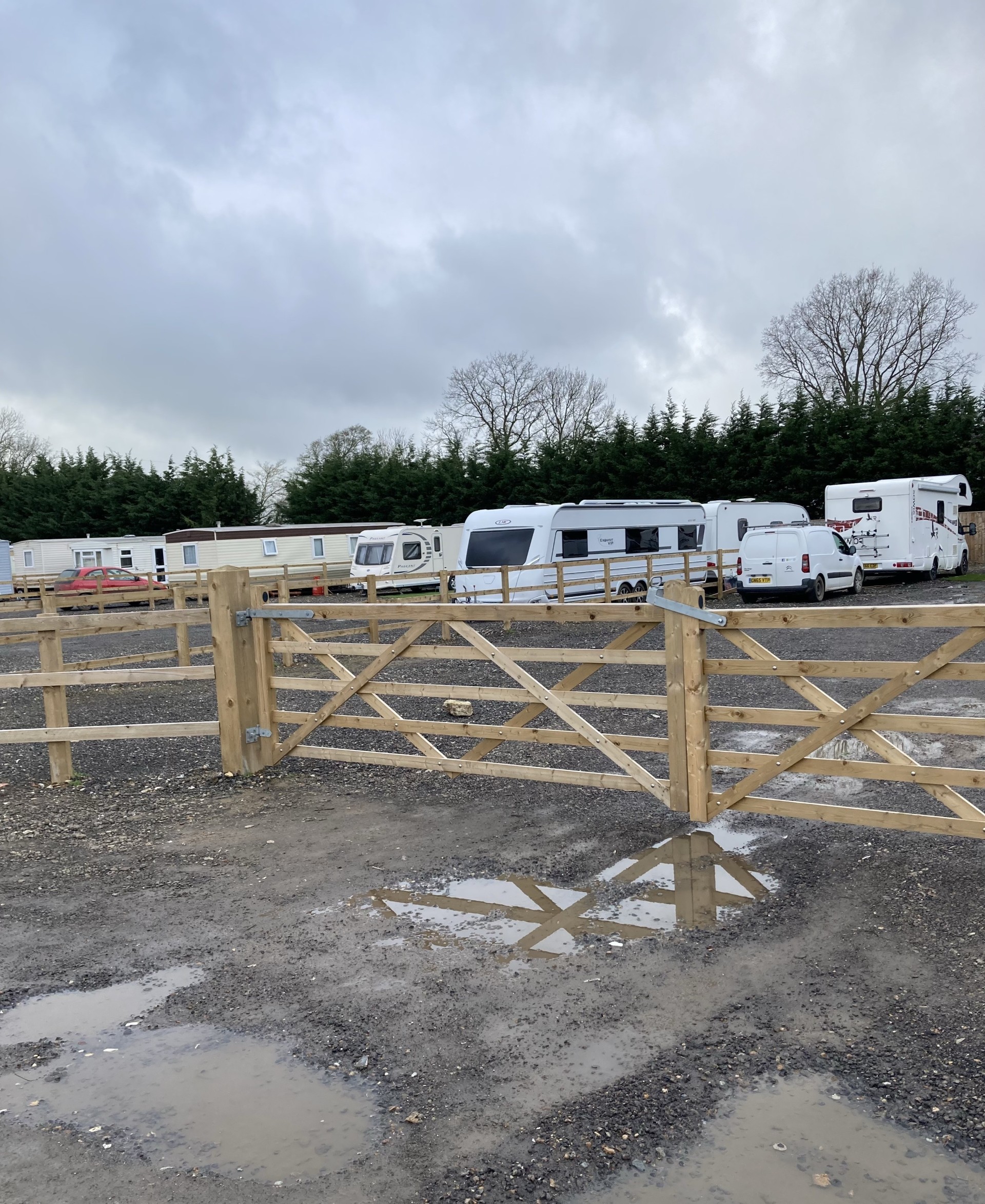 Muddy land with a brown fence. Behind the fence is a number of caravans and vehicles.