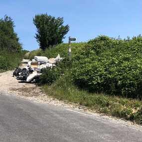 Country lane surrounded by green hedges, blue skies and pile of rubbish waste dumped on the roadway.