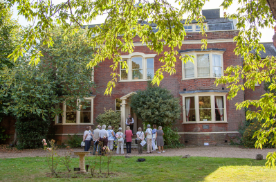 People standing in the doorway to a red bricked home, set in a beautiful green garden. Gads Hill is the former home to Charles Dickens.