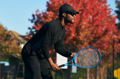 Man wearing black and a black hat holding tennis racket in position ready to serve